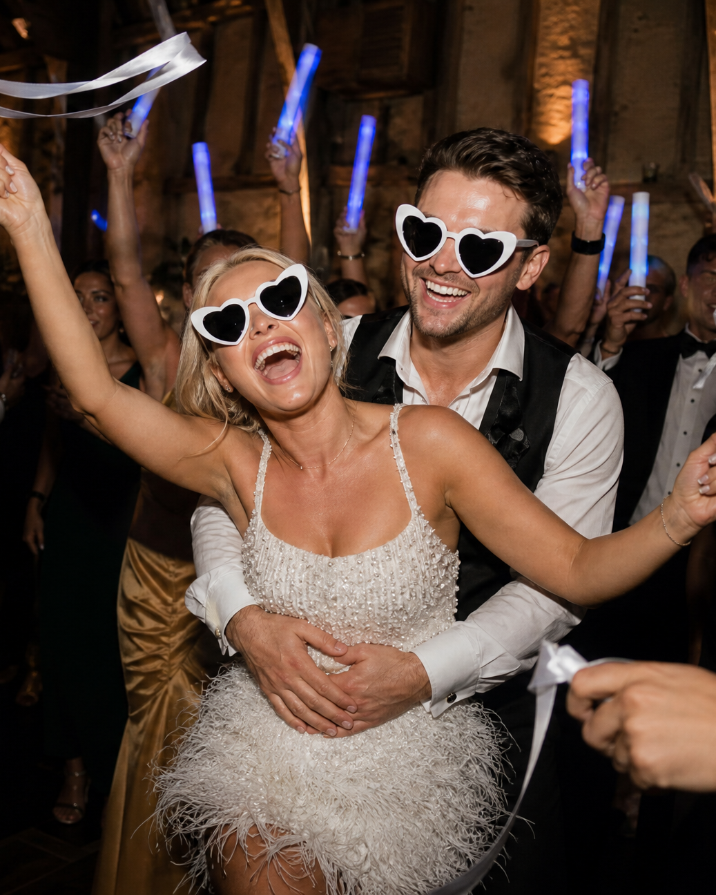 Late on the dance floor in the barn: the bride laughing with her arms up and the groom hugging her from behind, both in heart-shaped sunglasses, ribbon wands and glow sticks all around